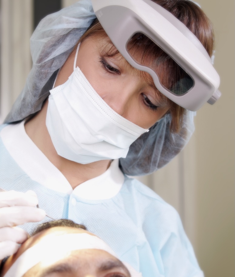 Dr Mari Carmen Morales performing a procedure on a patient. She is wearing a mask, surgical clothing, gloves, hair net, and has magnifying glasses on her forhead. The patient is a male who is leaning back and receiving a treatment on his scalp.